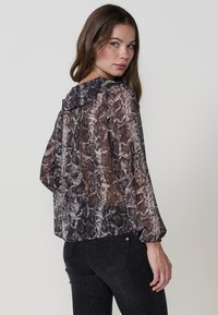 Blusa trasparente a maniche lunghe con motivo serpente nei toni di nero, bianco e marrone. Presenta un colletto arricciato e maniche voluminose.