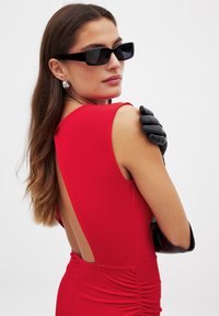 Czerwona sukienka bez rękawów z wycięciem na plecach, o marszczonej fakturze. Czarny rękawiczki i okulary przeciwsłoneczne dopełniają stylizację. Minimalistyczny design.