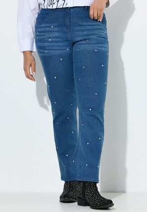 Niebieskie jeansy z denimu ozdobione rozrzuconymi małymi srebrnymi ćwiekami, noszone z czarnymi botkami na kostkę z białymi plamkami i niskim obcasem.