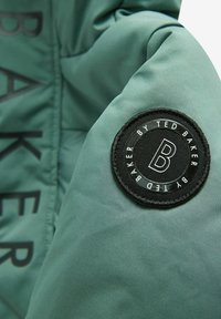 Roheline puhvjakki varrukas, millel on ümmargune must laik, millel on kiri "BY TED BAKER" ja täht "B", sile tekstuur ja matte viimistlus.