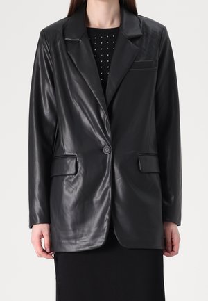 Kvinde iført en sort enkeltknappet faux læderblazer over en sort kjole med små firkantede pynt ved halsudskæringen.