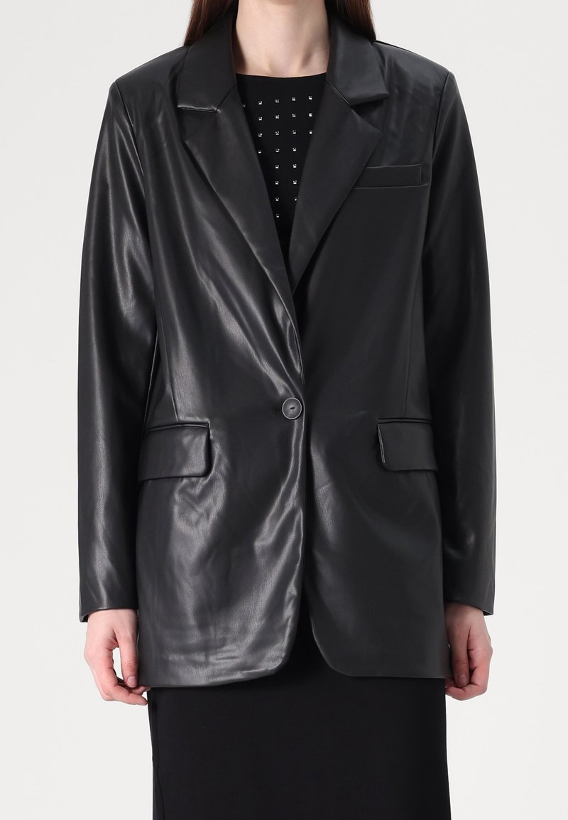 Femme portant un blazer en faux cuir noir à un bouton sur une robe noire avec de petites décorations carrées au niveau du col.