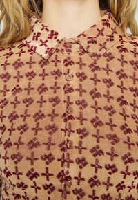 Tan korduroy košile s límcem, která má vzor v barvě burgundy se srdci a kříži. Šest knoflíků vpředu s texturovaným povrchem.