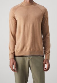 Beiger Rundhalsausschnitt-Pullover mit langen Ärmeln, gerippten Bündchen und Saum, der eine subtile Textur aufweist. Trägt er über olivgrünen Hosen.