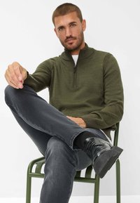 Pull à col zip olive, en tricot texturé, associé à un jean gris foncé et des bottes à lacets noires, assis sur une chaise verte devant un fond blanc.