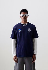Camiseta azul marino de manga corta con logo en el pecho y camiseta de manga larga blanca por debajo. Gafas de sol plateadas que reflejan la luz. Jeans grises completan el conjunto.