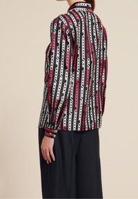 Schwarze Bluse mit vertikalem Kettenmuster in Weiß und Rot, mit Kragen und langen, leicht gepufften Ärmeln. Figurbetonte Passform.