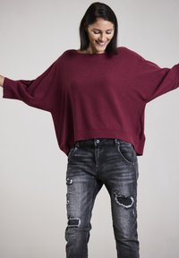Burgunderfarbener Oversized-Pullover mit Fledermausärmeln, kombiniert mit lädierten schwarzen Jeans, die sichtbare Risse und eine schmale Passform aufweisen.