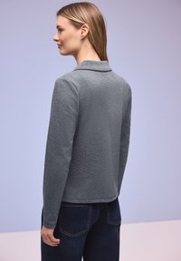 Femme aux cheveux blonds portant un haut gris à manches longues et un jean foncé, debout et regardant vers la gauche sur un fond uni.