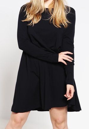 Blondynka ubrana w luźną czarną sukienkę z długim rękawem, stojąca na tle jednolitego jasnego tła.