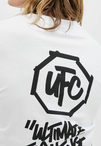 Sort UFC-logo og stiliseret "Ultimate"-tekst trykt på bagsiden af en hvid skjorte, båret af en person med lysebrunt hår.