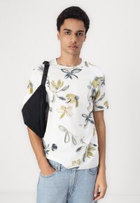 Biela tričko s krátkym rukávom s kvetinovým vzorom v zelených a žltých odtieňoch, skombinované so svetlomodrými džínsami a černou crossbody taškou.