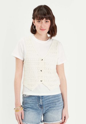 Gilet en crochet crème avec un col en V, présentant des boutonnières et un ourlet pointu, superposé sur un t-shirt blanc à manches courtes.