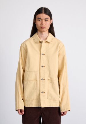 Veste en coton jaune avec un col, des boutons sur le devant et deux grandes poches avant. Présente des coutures visibles et un détail de logo subtil.