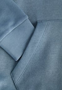 Blåt sweatstof med tekstureret overflade, ribstrikkede manchetter og en kængurulomme. Sømlyd er synlig nær lommen.