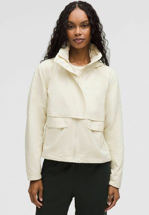Veste légère de couleur crème avec un col montant, fermeture éclair à l'avant et deux grandes poches. Présente un design court et un tissu texturé.