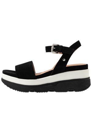 Sandalo in suede nero con una larga fascia, chiusura alla caviglia e una suola piatta a strati bianca e nera. Suola testurizzata per una migliore aderenza.