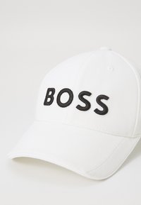 Fehér, ívelt karimájú vászon sapka; az elején fekete hímzett "BOSS" felirattal. Simább textúra, strukturált kialakítás.