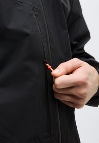 Main tenant un curseur de fermeture éclair orange sur une veste noire, gros plan sur le torse et la main sur un fond uni clair.