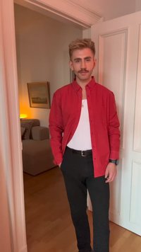 Jeune homme avec une moustache portant une chemise rouge sur un haut blanc et un pantalon noir se tenant dans une porte à l'intérieur d'une pièce faiblement éclairée.