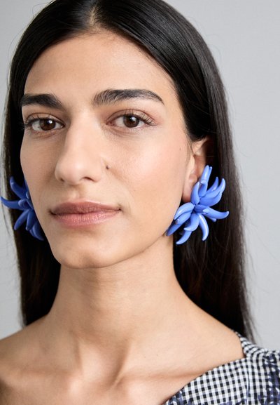 Cercei în formă de floare, de culoare albastră, din plastic, cu petale alungite și o textură netedă, asortați cu un top în carouri.