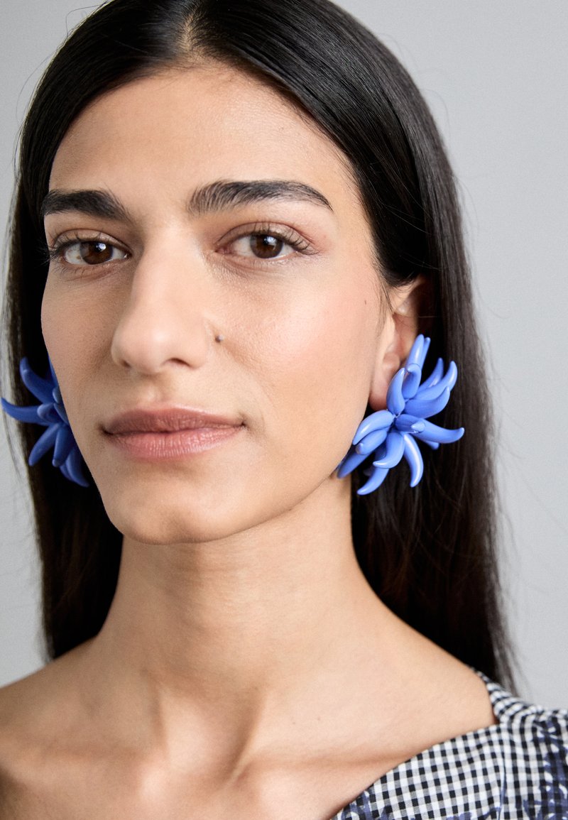 Pendientes azules en forma de flor, hechos de plástico, con pétalos alargados y una textura suave, combinados con una blusa de cuadros.