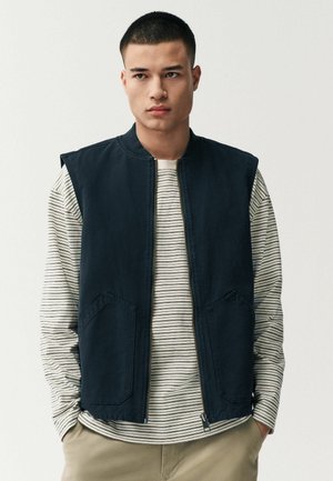 Navy vest with a round neckline, front zipper, and two large pockets, worn over a white and gray striped long-sleeve shirt.