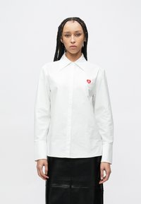 Bijela pamučna košulja na kopčanje s oštrim ovratnikom, s malim crvenim logotipom srca na lijevoj strani prsa. Duge rukave i ravni kroj.