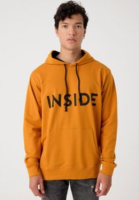 Sudadera naranja con un gráfico negro "INSIDE", cordones ajustables, bolsillo frontal y puños de canalé, llevada sobre una camiseta negra.