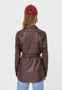 Brązowa koszula z ekoskóry z paskiem, długimi rękawami, mankietami na guziki i detalem na plecach, zestawiona z jasnoniebieskimi dżinsami i czerwoną czapką.