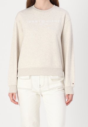 Femme portant un sweat-shirt beige Tommy Hilfiger avec logo et un pantalon clair, debout devant un fond blanc uni.