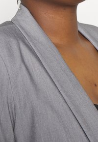 Grauer Blazer mit glatter Textur und breiten Revers, versehen mit einem dezenten Glanz und taillierter Passform, gezeigt von der Schulter und dem Ausschnitt.