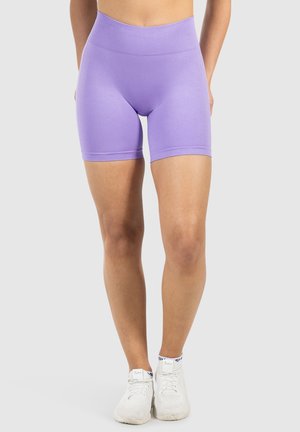Een persoon staat gekleed in paarse sportshorts en witte sneakers, en toont een sportieve outfit tegen een effen achtergrond.