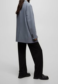 Blazer gris clair avec un col cranté, une fente arrière simple et des boutons noirs, associé à un pantalon noir ample et des chaussures noires épaisses.