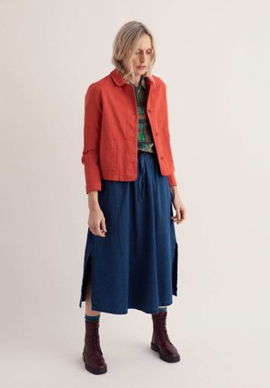 Frau mit blonden Haaren, die eine orange Jacke, ein grün kariertes Hemd, einen blauen Midi-Rock, bordeauxrote Stiefel und türkisfarbene Socken trägt, steht vor einem schlichten Hintergrund.
