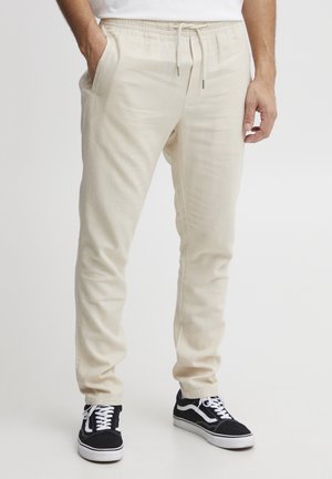 Homme portant un pantalon jogger écru avec cordon et des baskets noir et blanc, debout, une main dans la poche, sur un fond uni.