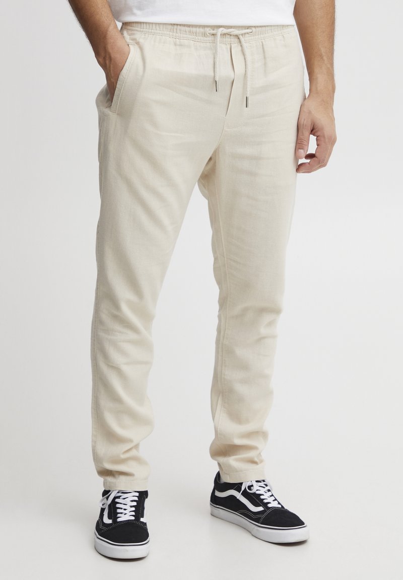 Homme portant un pantalon jogger écru avec cordon et des baskets noir et blanc, debout, une main dans la poche, sur un fond uni.