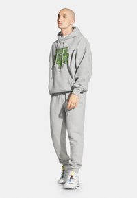 Szara bluza z kapturem z zielonym napisem graficznym, dopasowane spodnie dresowe i białe sneakersy. Materiał wydaje się być miękki; luźny krój z kapturem na sznurki.