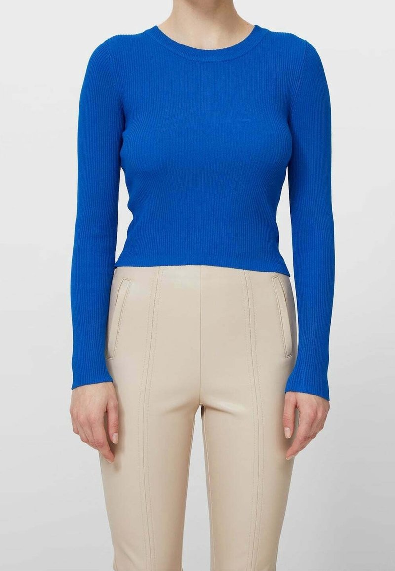 Femme portant un haut bleu côtelé ajusté à manches longues et un pantalon beige taille haute avec des détails de coutures devant et des poches.