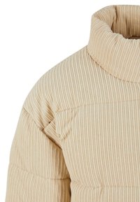 Béžová prošívaná bunda s žebrovaným vzorem, vysokým límcem a prošitým designem, představující měkkou texturovanou látku s vertikálními pruhy.