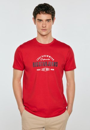 Młody mężczyzna w czerwonej koszulce z nadrukiem z napisem "BIG STAR JEANS" oraz beżowych spodniach, stojący z rękami w kieszeniach na jednolitym tle.