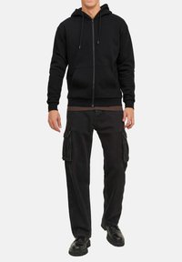 Schwarze Zip-Hoodie mit Kordelzügen, kombiniert mit lockeren schwarzen Cargo-Hosen mit Seitentaschen und robusten schwarzen Schuhen.