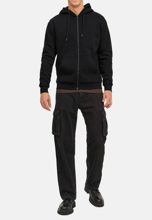 Zwarte zip-up hoodie met trekkoorden, gecombineerd met los fitting zwarte cargo broek met zijzakken en stevige zwarte schoenen.