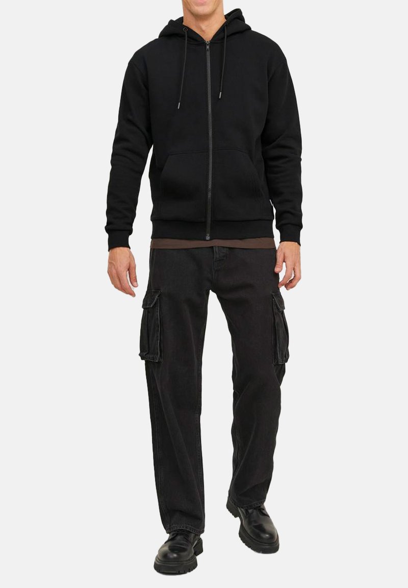 Schwarze Zip-Hoodie mit Kordelzügen, kombiniert mit lockeren schwarzen Cargo-Hosen mit Seitentaschen und robusten schwarzen Schuhen.