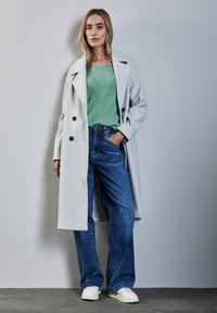 Lichtgrijze wolmixjas met dubbele knopen, groene gebreide trui, blauwe jeans en witte sneakers op een grijze achtergrond.