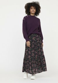 Pull en maille violet avec un motif texturé et des boutons dorés à l'épaule, associé à une longue jupe fleurie aux tons sombres et des baskets blanches.