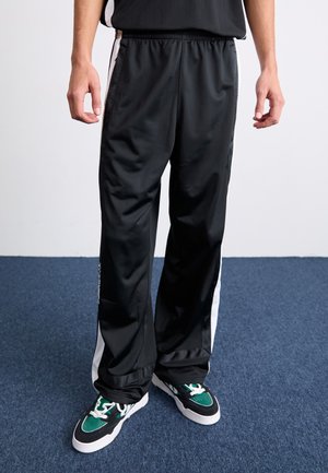 Person iført sorte træningsbukser med hvide striber og grønne, sorte og hvide sneakers, stående på blåt tæppe.