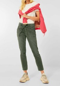 Olivově zelené kalhoty se stahováním v pase, bočními kapsami a úzkým střihem. Kombinovány se světle barevným tričkem a růžovou bundou.