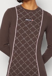 Brązowa bluzka z długim rękawem, ozdobiona powtarzającym się geometrycznym wzorem, różowymi akcentami w postaci lamówki oraz czarno-białą naszywką z logo "ellesse" z przodu.
