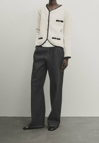 Persoon in een crèmekleurige textuurjacket met zwarte bies en knopen, brede donkere jeans en zwarte schoenen, staande tegen een effen muur.